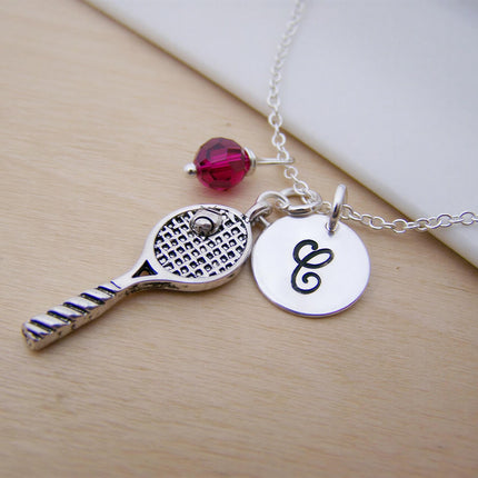 Silver tennis racquet necklace with a pink bead and a round charm printed with the letter C on a thin metal chain.
