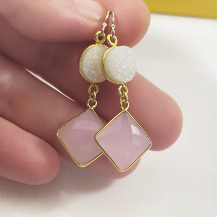 Two gold earrings with round white druzy studs and square pink gemstone dangles held between two fingers.